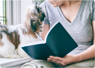 Mulher tomando notas do curso educacional em um caderno enquanto olha para seu cachorro ao lado dela.
